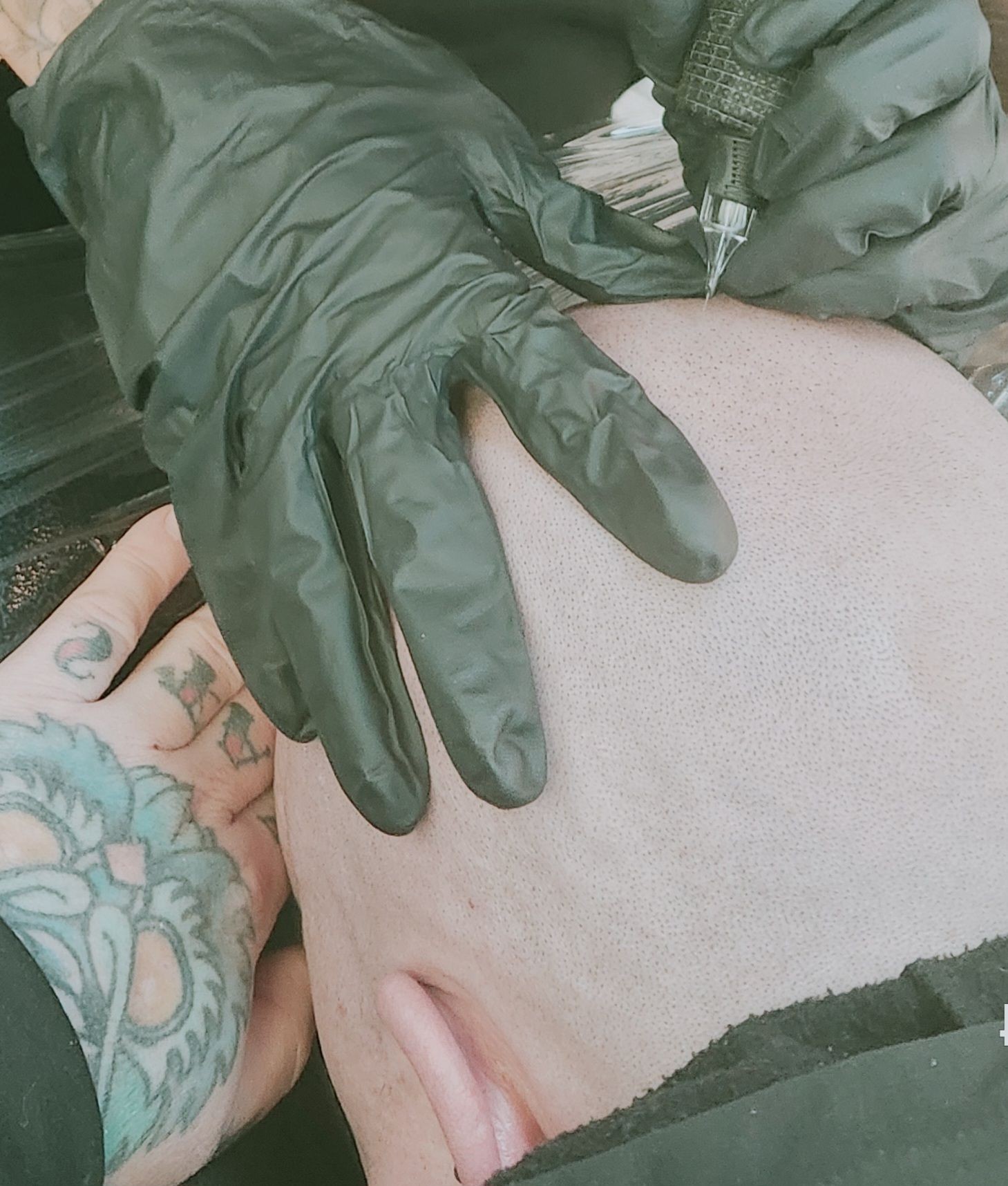 Artist wearing black gloves tattooing a design on the back of a person's head.